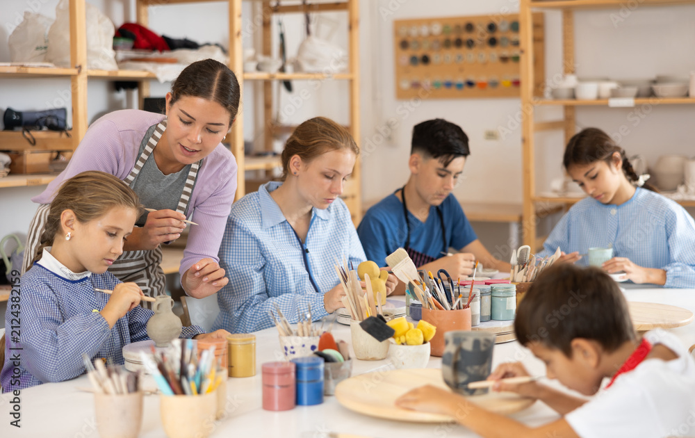 Fototapeta premium Teenagers and children at a ceramics workshop in an art studio. Woman teacher shows how to paint clay dishes with a brush