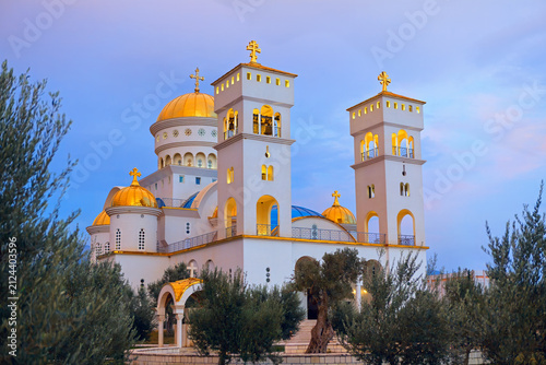 Church of St Jovan Vladimir stands tall in Bar, Montenegro under moody twilight sky. Impressive Orthodox cathedral features gleaming golden domes, white stone walls, and surrounding olive trees
