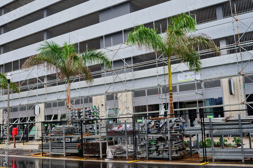 Scaffolding and construction materials line a downtown building frontage flanked by palm trees, wet pavement and reflective puddles after rain, conveying urban renovation and temporary disruption.
