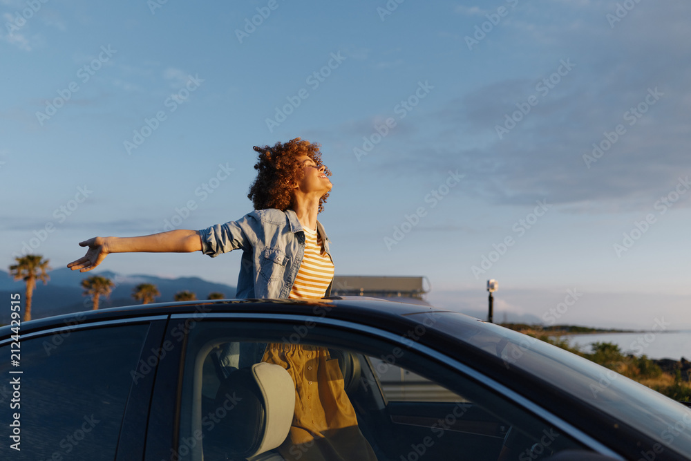 Fototapeta premium A woman with curly hair stands through an open car window along a sunny coastline, arms outstretched. A lifestyle moment of freedom, travel, and optimistic daydreams at the seaside.