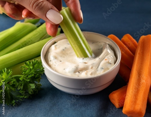 hand dipping celery sticks in ranch dip with a bowl of carrots and a background