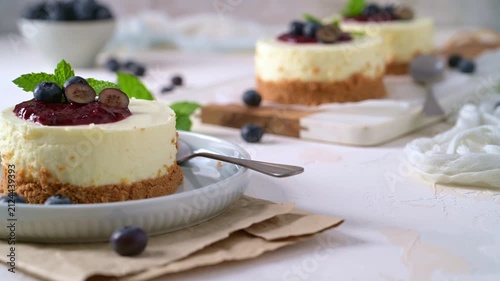 Mini cheesecakes topped with vibrant berry compote and fresh mint are illuminated by soft natural light on a rustic white background with scattered blueberries