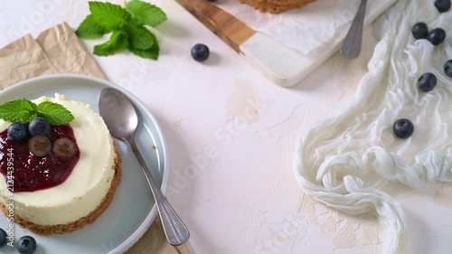 Mini cheesecake topped with berry compote and fresh mint, illuminated by soft natural light on a textured white background with scattered berries.