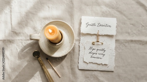 Serene lit beeswax candle sits on ceramic holder beside vintage brass wick trimmer and handmade paper with inspirational quote text on linen background under natural sunlight for calm home brandings.