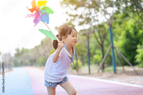 Obraz na plátně Cheerful little girl running forward while holding a colorful pinwheel toy on a park path