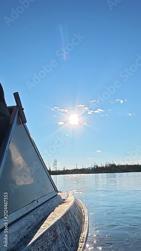 A serene view from a boat on a calm lake with bright sunlight shining through scattered clouds in a clear blue sky.