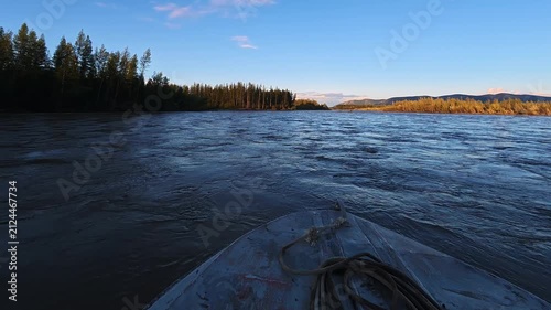 Front view from a small boat navigating calm river waters, surrounded by dense forests and rolling hills under a clear blue sky during golden hour.