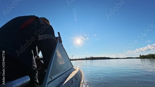 A person in dark attire operates a small boat on a calm lake under a bright sunny sky, with the sun casting rays over the water and distant forested shoreline.