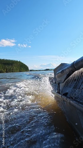 A motorboat travels along a wide river, creating a frothy wake as it moves past dense green forests with distant snow-capped mountains visible under a clear blue sky.