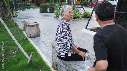 Adult Asian son caring for elderly mother sitting outdoors in park. Intergenerational family relationship and home caregiver concept.