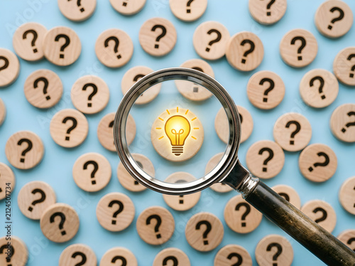 Magnifying glass focusing on a glowing light bulb symbol surrounded by many wooden question marks with question mark solution discovery innovation creativity thinking knowledge business