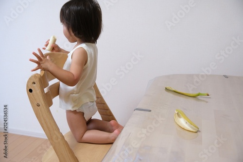 Toddler wearing diaper holding a banana and standing on a wooden chair