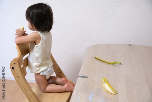 Child wearing diaper standing on a wooden chair and eating a banana