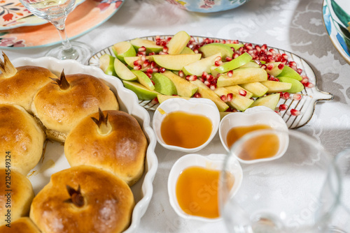 A festive table setting shows round challah bread and sliced apples with honey on the side. There are pomegranate seeds on the apples. This is a part of Rosh Hashanah traditions.