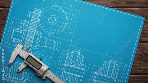 Digital vernier caliper placed on mechanical engineering blueprints with gear drawings on a dark wooden background.