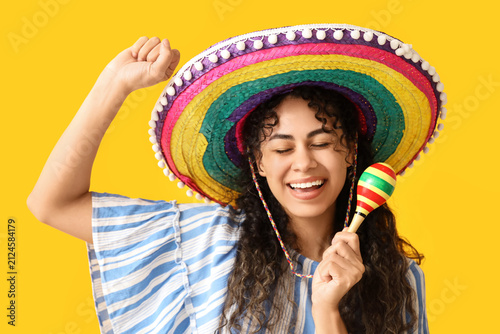Fotografie Beautiful young African-American woman in sombrero hat with maracas on yellow ba