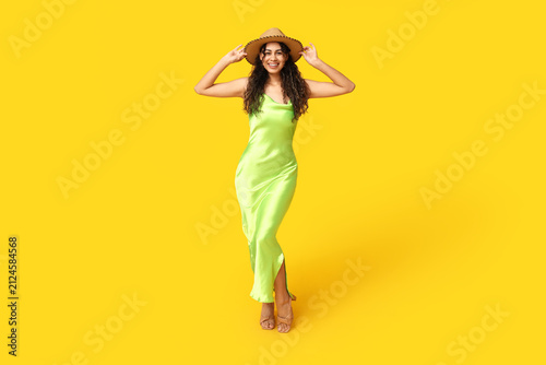 Stylish African-American woman in dress and hat on yellow background