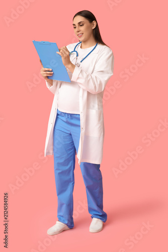 Beautiful young pregnant doctor with clipboard on pink background