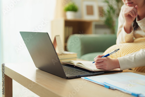 Fotografie Young woman taking notes while studying online with laptop at home