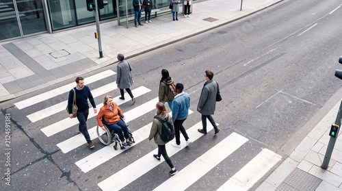 Pedestrians Crossing Urban Street: A diverse group of people, including a person in a wheelchair, navigate a busy city street at a crosswalk, showcasing urban life and accessibility.