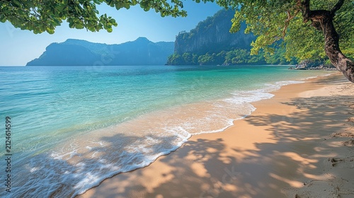 Clear turquoise sea waves gently washing golden sandy tropical beach with tree shadows and lush green mountains under a blue sky.