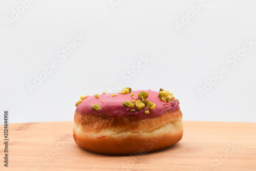 A close-up of a single donut topped with pink glaze and crushed pistachios, sitting on a wooden board against a clean white background, evoking fresh bakery flavors and sweet indulgence.