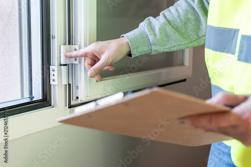 Property inspector wearing a reflective vest checking the condition and functionality of a modern window frame, holding a clipboard to document findings during a real estate assessment