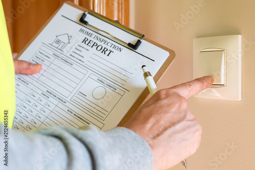 Professional home inspector documenting property condition and safety issues, examining an electrical light switch while holding a clipboard with a home inspection report form