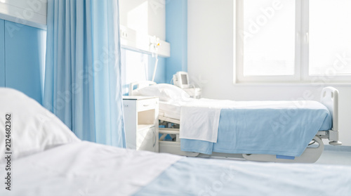 Empty hospital bed covered with blue blankets positioned next to privacy curtains inside bright clinic room featuring medical monitoring equipment near large window