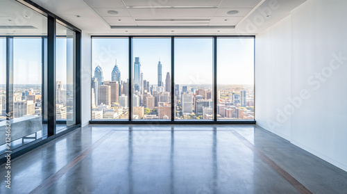 Empty open plan corporate workspace interior featuring wooden desks equipped with computer monitors resting on concrete floors alongside panoramic city skyline windows