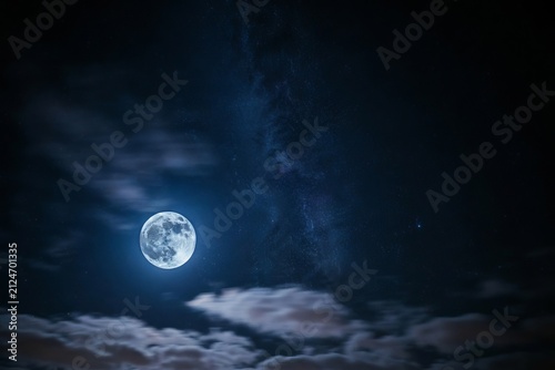 A bright full moon glowing in the dark night sky surrounded by scattered clouds and the faint hazy band of the milky way galaxy visible above a dark horizon in a serene nocturnal landscape scene