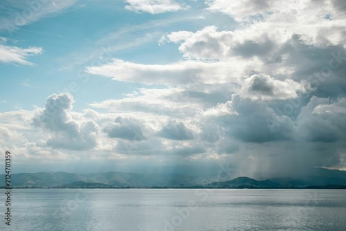 A calm lake reflecting a dramatic sky filled with heavy grey rain clouds and patches of blue while distant mountains sit on the horizon under a light shower falling over the water surface area