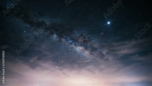 The bright band of the milky way galaxy stretching across a dark night sky filled with countless stars and faint clouds above a glowing horizon line in a deep space astronomical scene