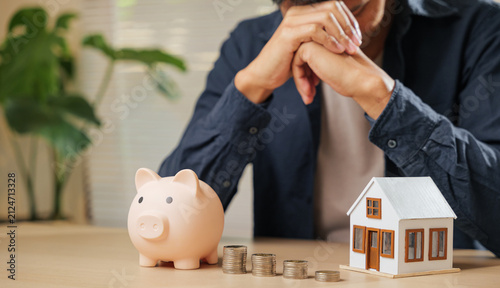 Cuadro en lienzo Stressed man holding his forehead, surrounded by a piggy bank, coin stacks, and a house model