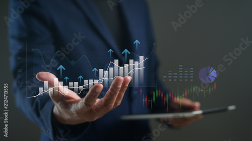 A businessperson in a suit holds a tablet displaying digital financial graphs and charts with rising arrows, symbolizing growth and data analytics.