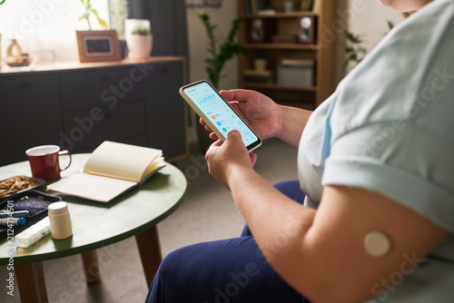 Caucasian woman with diabetes checking glucose levels on smartphone app, wearing continuous glucose monitor sensor on arm. Useful for digital health and telemedicine marketing