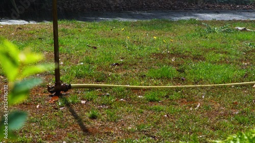 A tall commercial-grade sprinkler sprays a heavy volume of water across a lush, healthy lawn in a large garden under bright summer sunlight, surrounded by dense green foliage.