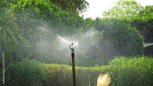 A tall commercial-grade sprinkler sprays a heavy volume of water across a lush, healthy lawn in a large garden under bright summer sunlight, surrounded by dense green foliage.
