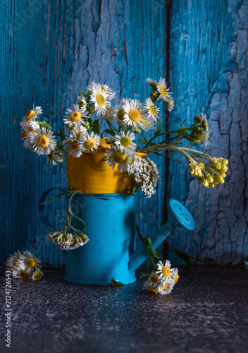 A small bouquet of meadow daisies in a blue watering can on a dark background.