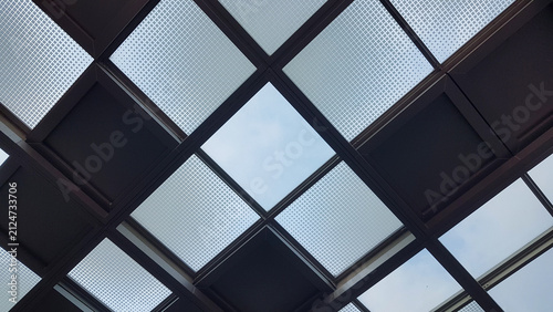 Low angle view looking up at a modern architectural skylight roof featuring a geometric grid pattern of dark metal frames and textured glass panels.