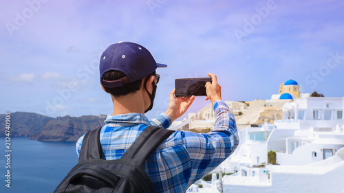 Tourist asian man taking photo with smartphone of famous blue domed churches in Oia village, Santorini island, Greece