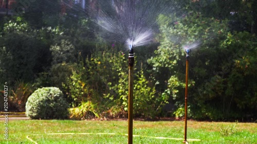 A tall commercial-grade sprinkler sprays a heavy volume of water across a lush, healthy lawn in a large garden under bright summer sunlight, surrounded by dense green foliage.