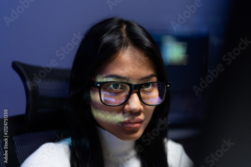 Portrait of Asian woman programmer with glasses looking at camera with code reflection on her face, responsible for checking AI generated code and fixing bugs at night