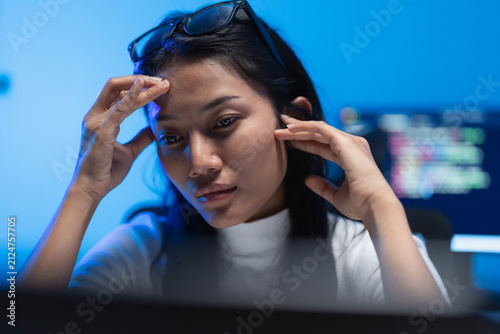 Woman software developer leans toward a screen with hands at her head, code visible on a monitor behind her in a blue lit room during a late night work shift.
