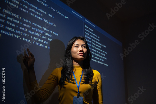 Woman software developer raises a finger as an idea comes to mind while reviewing projected code and server logs in a dark room, focused on solving a technical problem.
