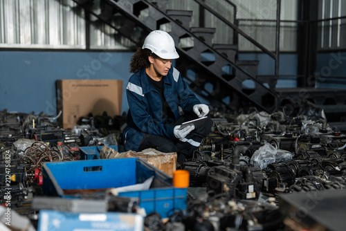 Diverse professional engineers in hard hats using a digital tablet for inventory management and inspecting machinery spare parts in a warehouse for e-waste recycling and sustainability.