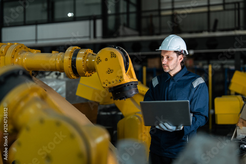 Professional male engineer in a blue uniform and hard hat using a laptop to inspect and program a yellow industrial robotic arm in a modern smart factory, focusing on industry 4.0 automation.
