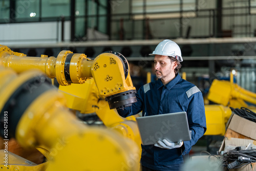 Professional male engineer in blue uniform and hard hat using a laptop to program and inspect a yellow industrial robotic arm in a modern smart factory for industry 4.0 automation.