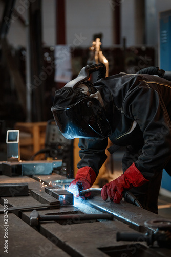 Fotografie Vertical image of young adult welder in protective mask and gloves welding metal