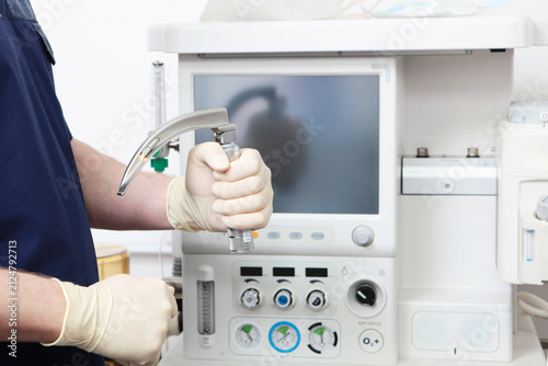 Doctor's hand in sterile gloves holds laryngoscope against background of modern anesthetic device in operating room